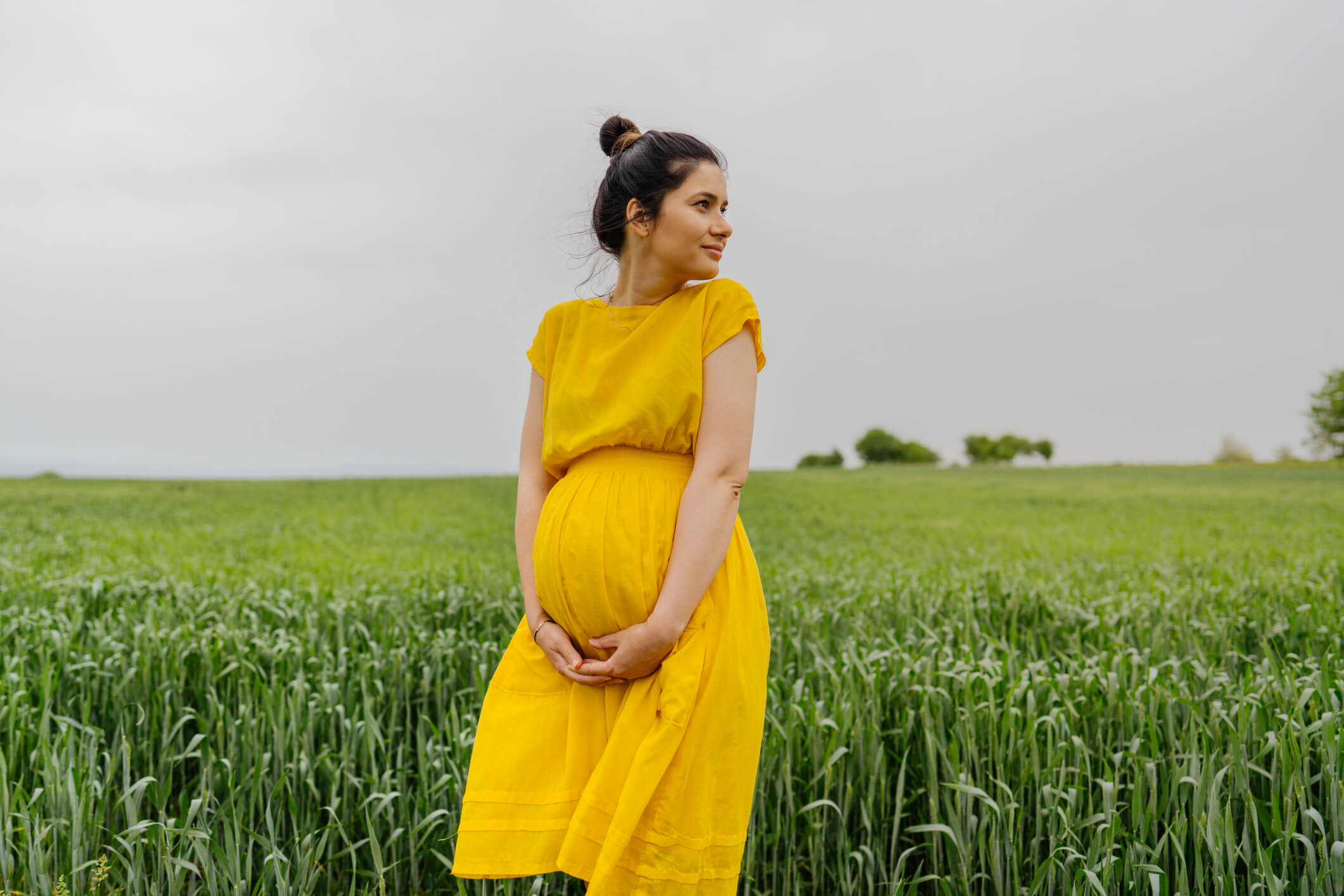 Photo of a pregnant woman standing alone on a grass field, stroking her baby bump and enjoying the calm meadow.